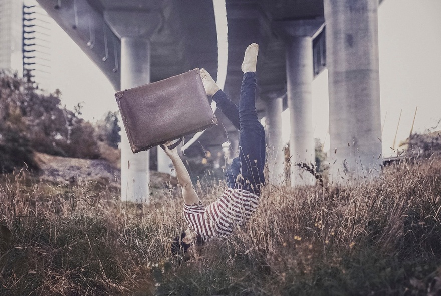 Self-portrait of an artist in a one-armed handstand, holding a suitcase with the other, underneath a large concrete bridge.