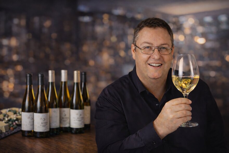 Man wearing a black shirt and glasses, holding a wineglass with 6 bottles behind him. 