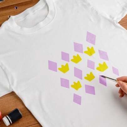 A hand holdign a small paintbrush working on decorating a t-shirt with purple diamonds and yellow crowns. 