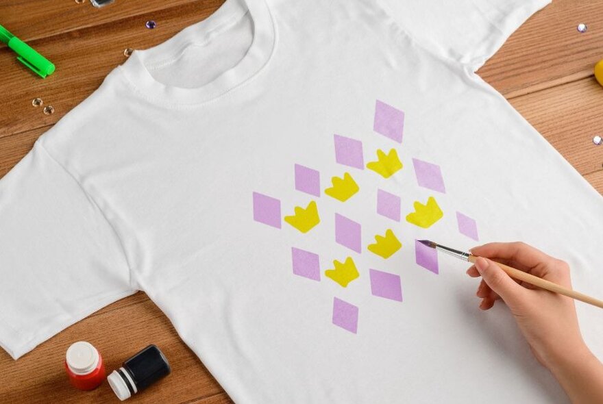 A hand holdign a small paintbrush working on decorating a t-shirt with purple diamonds and yellow crowns. 