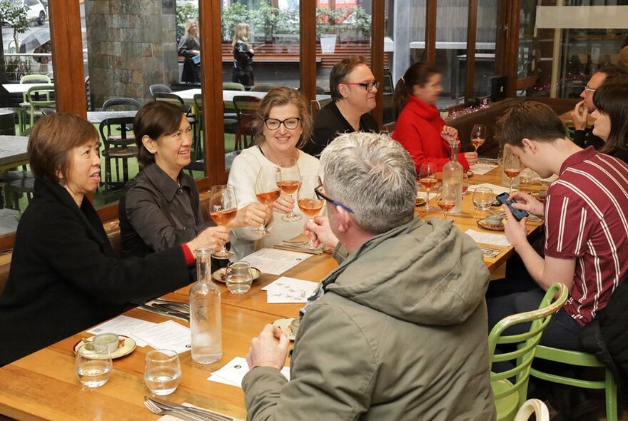 Dining patrons seated around a long table in a restaurant, all holding up their glasses of wine and bringing them together in a cheers gesture.
