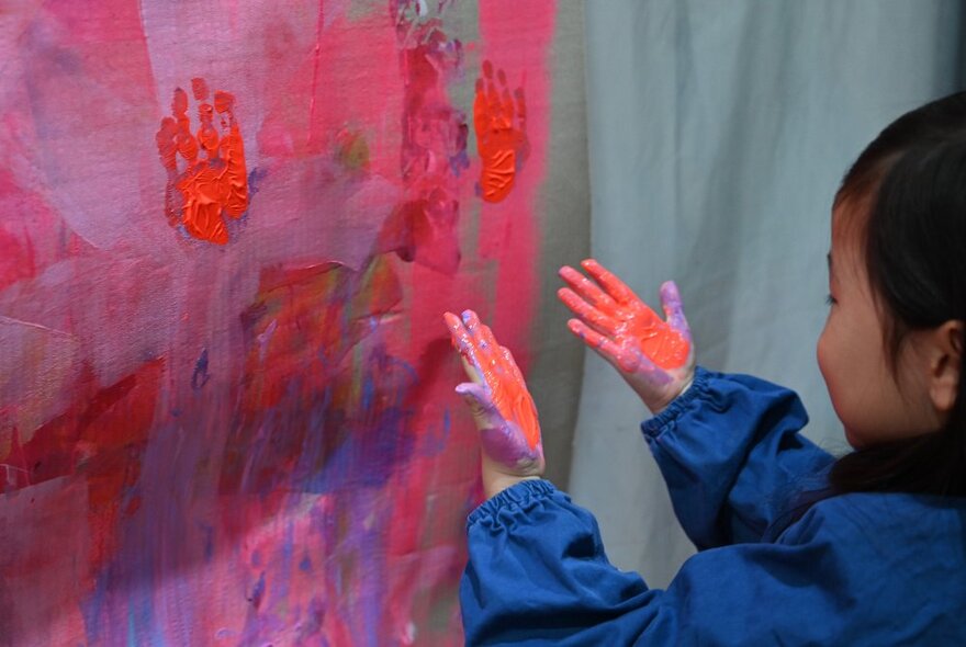 A child in an art smock in front of a painting, looking at their messy red painted hands