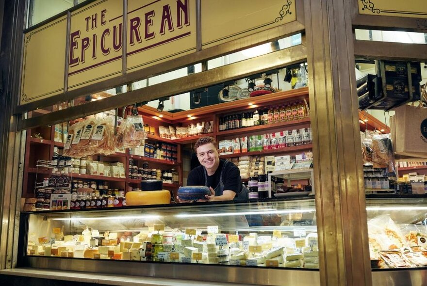 A shopkeeper at a cheese stall at the Queen Victorian Market, holding a wheel of cheese covered in blue wax over the counter.