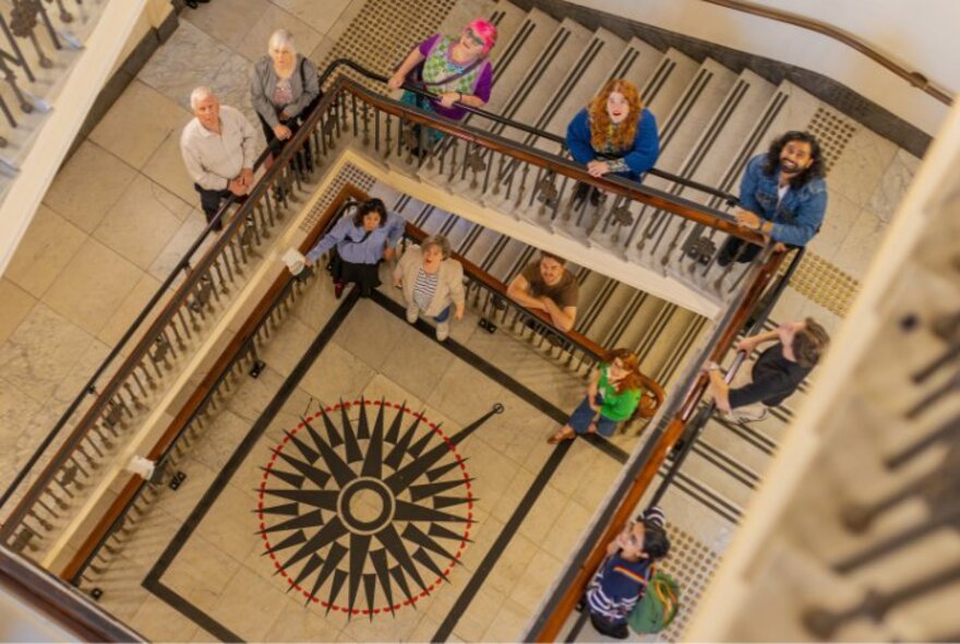 An overhead, high-angle shot looking down a multi-story, square-shaped historic stairwell with ornate iron railings and mosaic ground floor. Ten people are distributed across different landings and steps, looking up toward the camera.