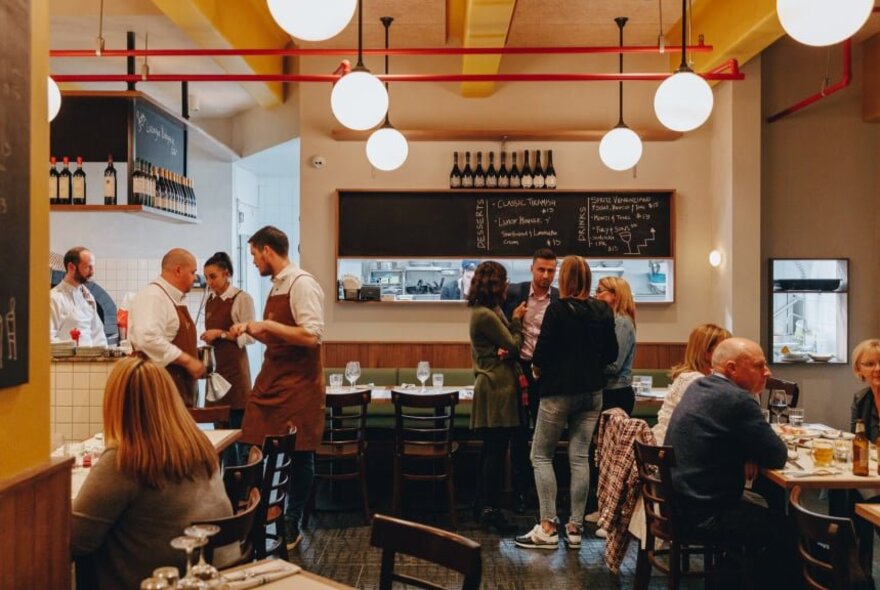 Interior of The Hardware Club restaurant, with a view into the kitchen, in the foreground are patrons dining, with wait staff standing in a huddle near the kitchen service area.