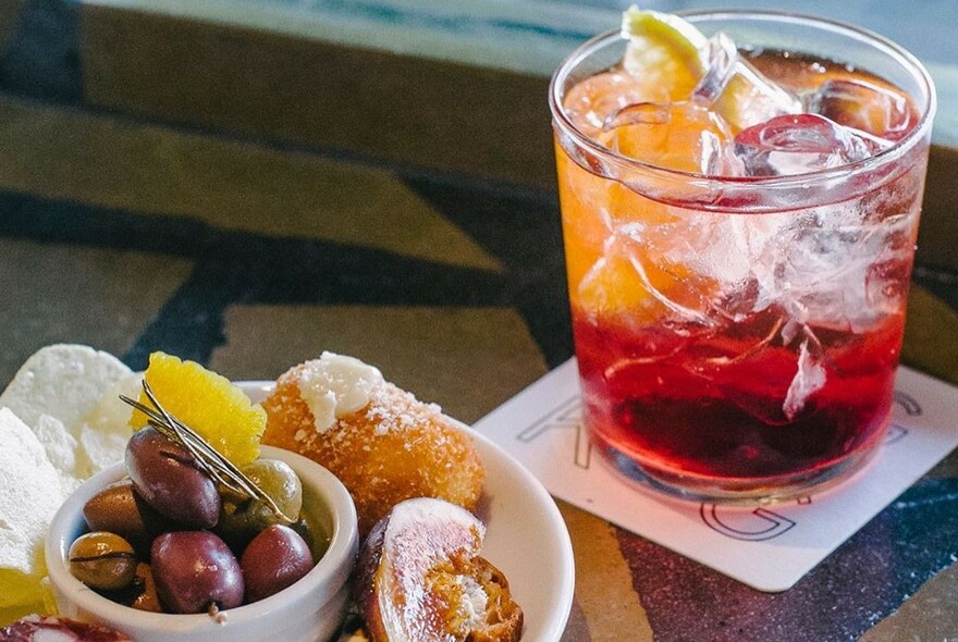 Iced red and orange-coloured drink with a small bowl of olives