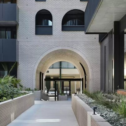 A wide outdoor stone walkway flanked by low brick planter boxes with green plants, leading toward a large arched opening in a modern white stone apartment building.