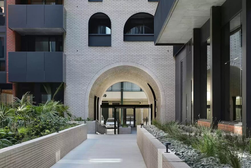 A wide outdoor stone walkway flanked by low brick planter boxes with green plants, leading toward a large arched opening in a modern white stone apartment building.