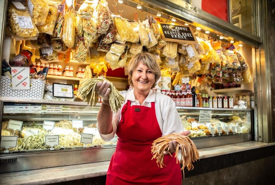 A woman in a red apron holding up handfuls of produce in front of a deli stall at the Queen Victoria Market. 