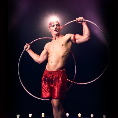 A circus performer in red satin shorts and a red bandana holding two silver hoops behind his back.
