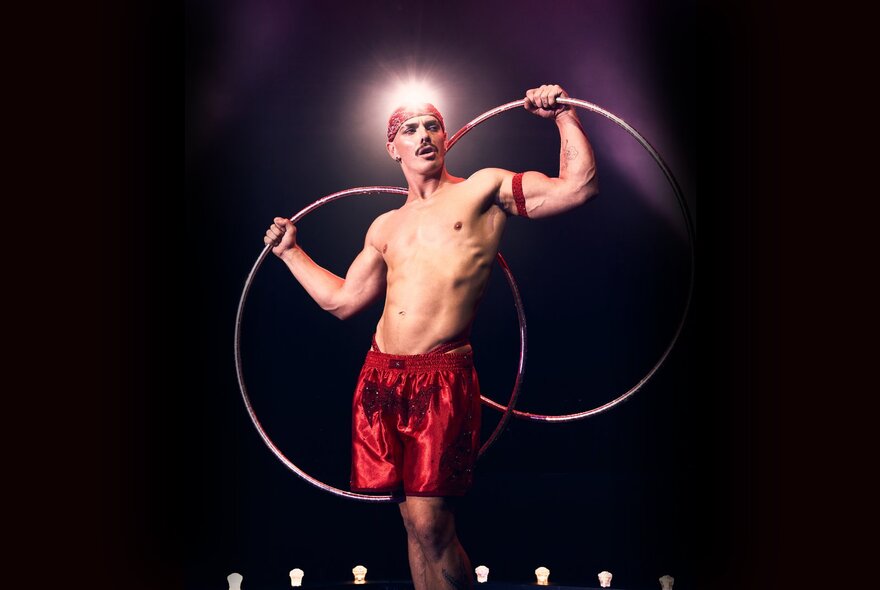 A circus performer in red satin shorts and a red bandana holding two silver hoops behind his back.