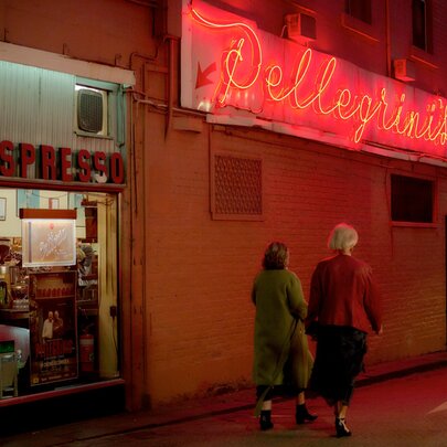 Two women walking under a neon sign at a corner cafe. 