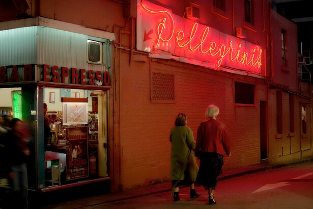 Two women walking under a neon sign at a corner cafe. 