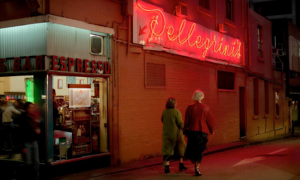Two women walking under a neon sign at a corner cafe. 