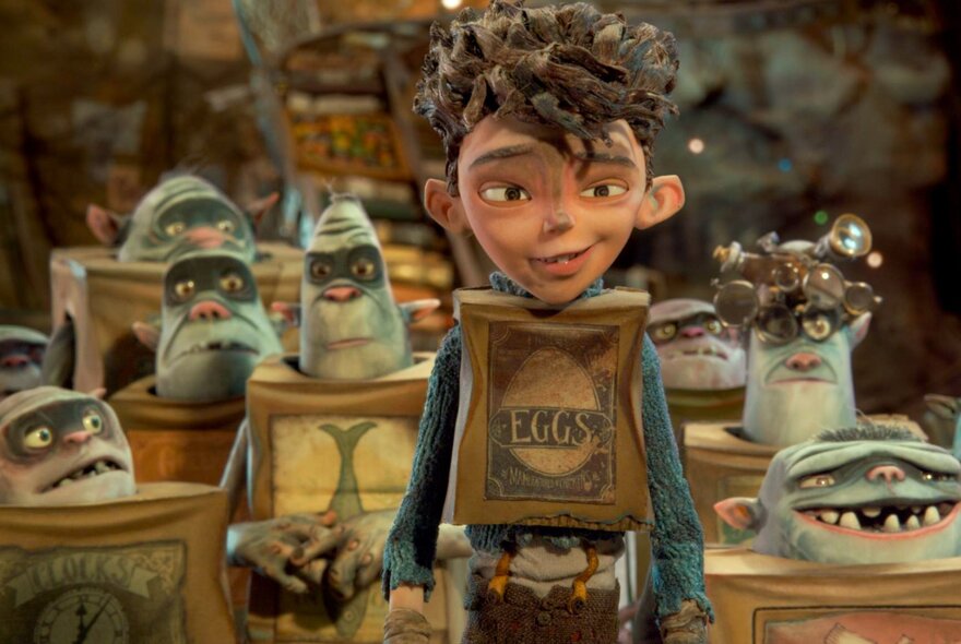 A still shot from the animated film The Boxtrolls, of a boy wearing a cardboard box labelled 'EGGS' smiling in the centre, surrounded by several grey troll creatures inside boxes in a dimly lit workshop.