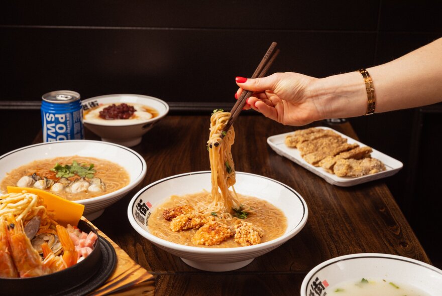 Hand using chopsticks to draw noodles out of a bowl of soup, with other Asian dishes on the table.