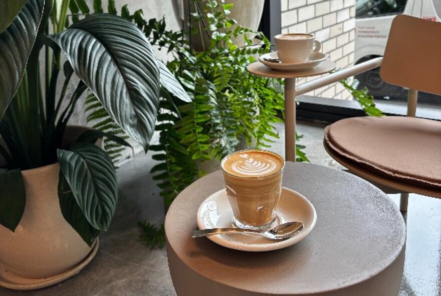 A glass of coffee on a small table in a cafe, with green pot plants amd a chair nearby.