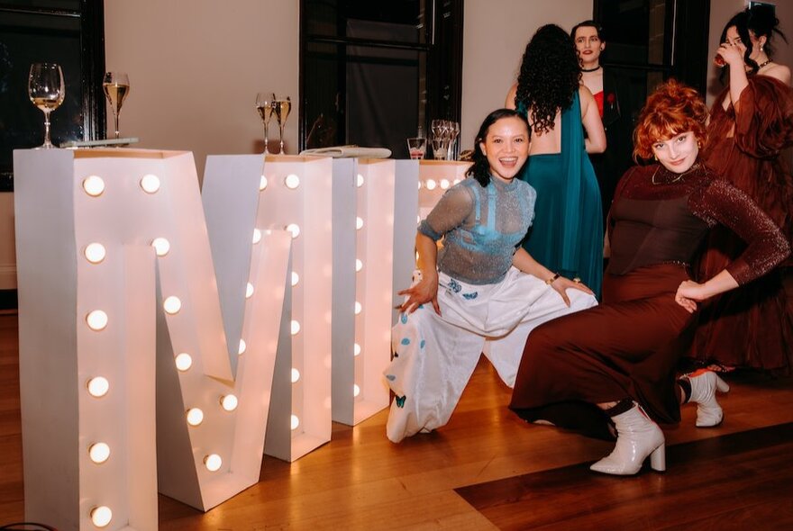 Two women posing next to large, white letters with lights inside them; the letters are M and I.
