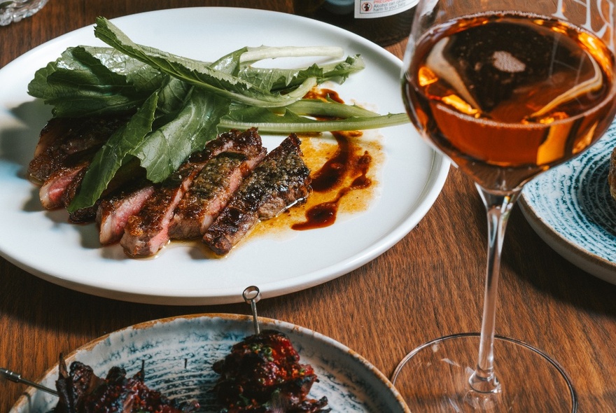 A table with a glass of wine and a main meal of sliced steak with greens. 