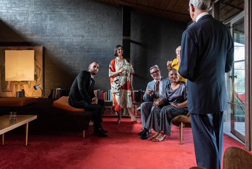 People seated in a modernist setting, listening to a man standing in front of them.