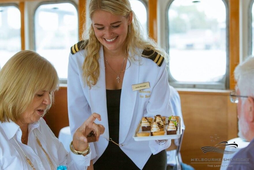 Waitress wearing a naval officer's jacket, serving petite desserts to customers inside a boat with windows looking onto the water.