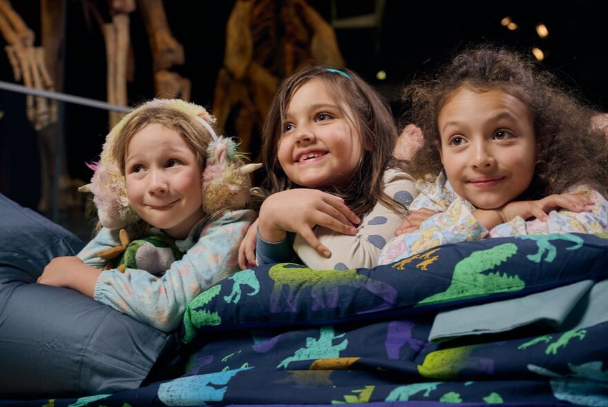 Three young girls on their tummies in a sleeping bag, one girl with fluffy earmuffs, under a dinosaur skeleton.