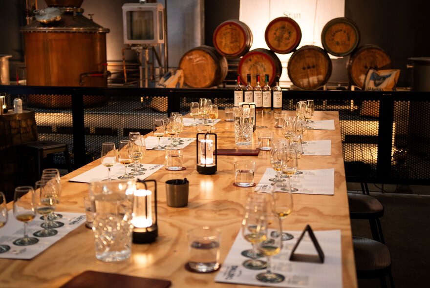 A long timber table with tasting glasses and candles, inside a whisky distillery, oak barrels in the background.
