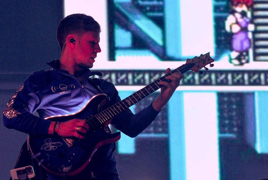 A man playing an electric guitar on a stage, with a large monitor in the background screening a retro game.