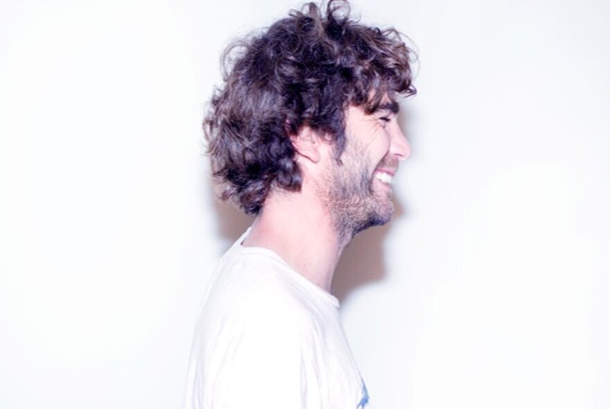 Man smiling in a mug shot, viewed from the side with curly dark hair, beard and white shirt.