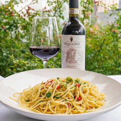 A white bowl of pasta with a bottle of red wine and a glass half-filled with wine behind the bowl, set against a green leafy view.