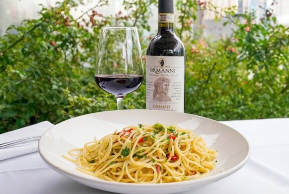 A white bowl of pasta with a bottle of red wine and a glass half-filled with wine behind the bowl, set against a green leafy view.