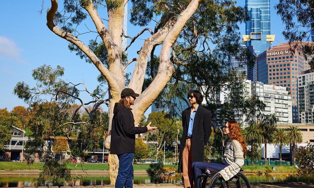Three people talking beside a large tree by a river in a city. One is using a wheelchair.