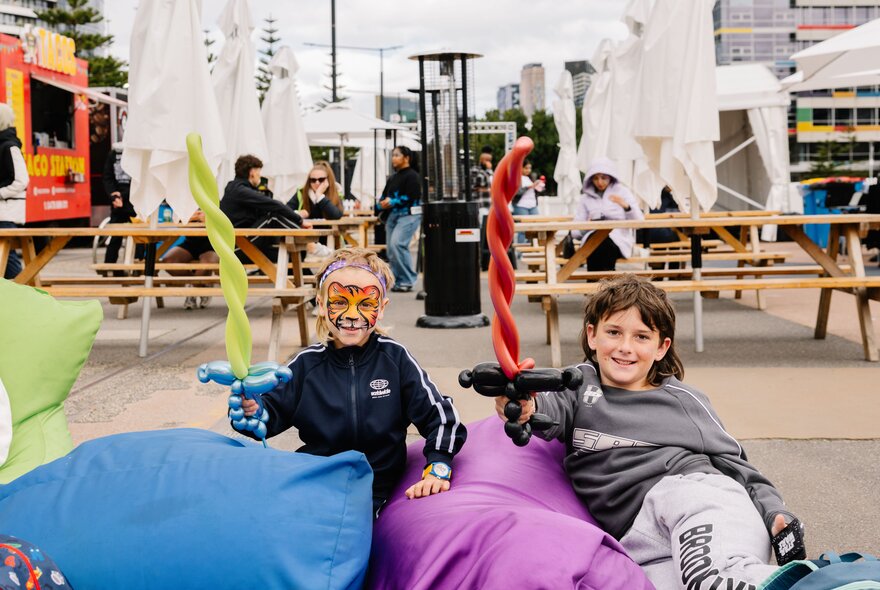 A family-friendly community pop-up event featuring children with face paint holding balloon swords while relaxing on outdoor beanbag chairs by the waterfront.