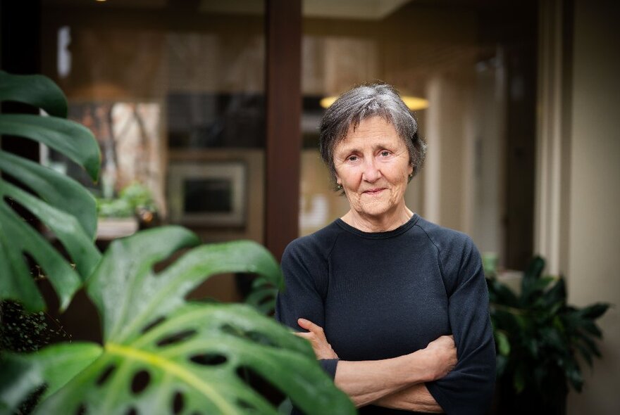 Author Helen Garner standing outside a room in a house, wearing a black top and with her arms crossed in front of her chest, looking directly ahead with a wry smile on her face.