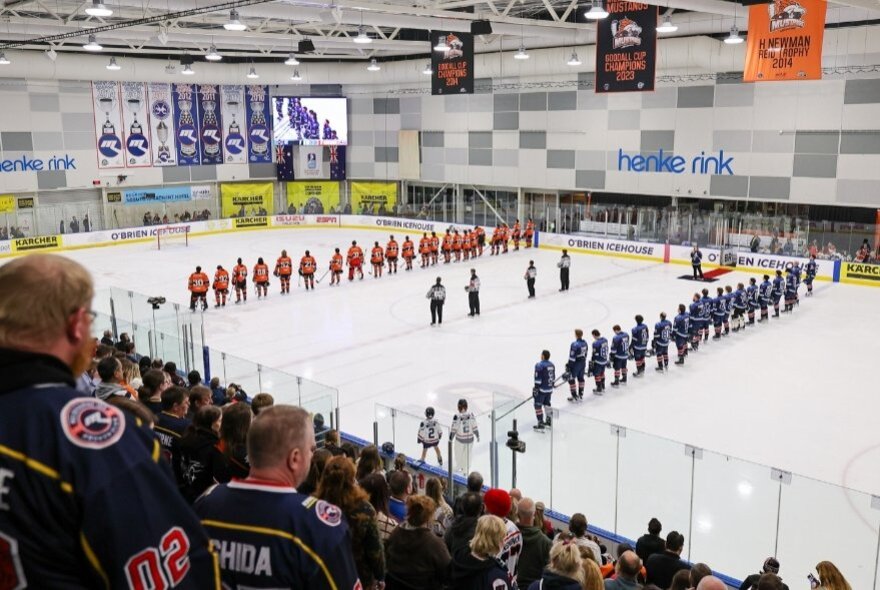 Two ice hockey teams lined up on the ice rink, facing each other, four referees in the middle.