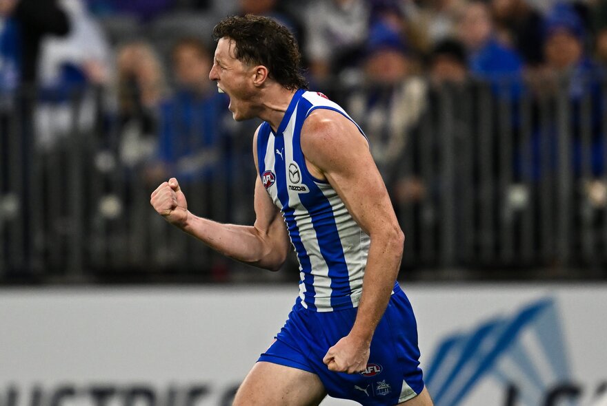 An AFL football player wearing the North Melbourne club kit, with his fist clenched and cheering, on the football field.