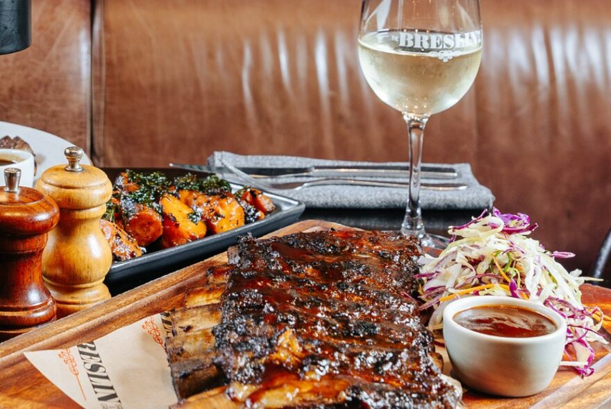 A plate of cooked meat on a wooden tray, on a dining table, with salt and pepper grinders and a glass of white wine.