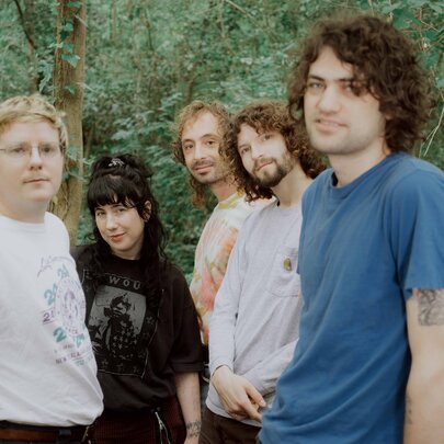 The five members of the indie rock band Wednesday, all looking at the camera, slightly smiling, standing outdoors in a forest setting.
