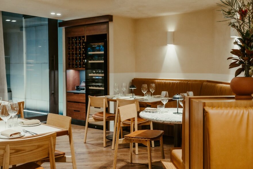 Interior of a softly lit restaurant with caramel coloured booth seating, and tables set for service with plates, glasses and cutlery.