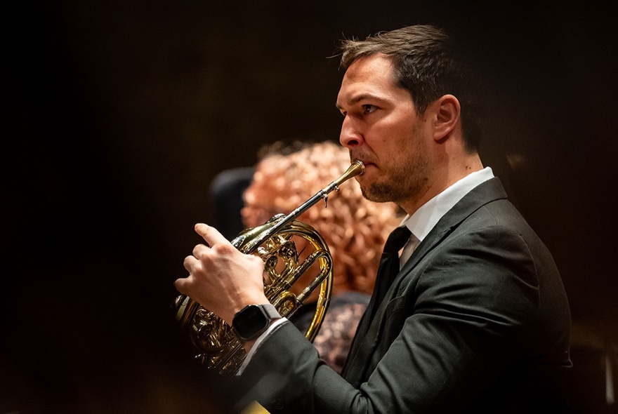 A man in a suit playing the French Horn in a concert setting. 