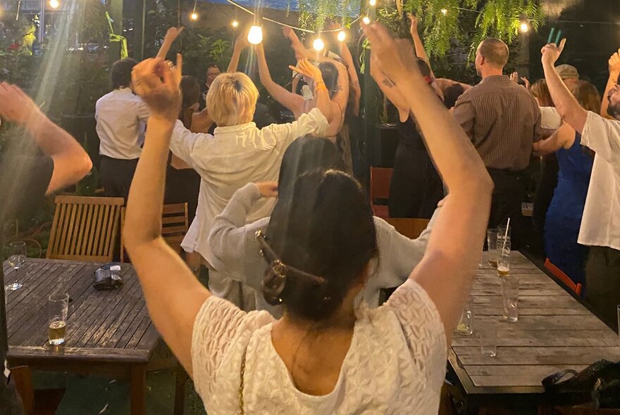 People seated and standing in a garden setting with outdoor lights, waving their arms in the air.