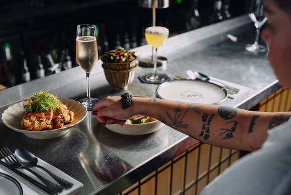A sleek stainless steel bar counter with drinks, small dishes, and a person's arm at Lexy Restaurant and Bar.