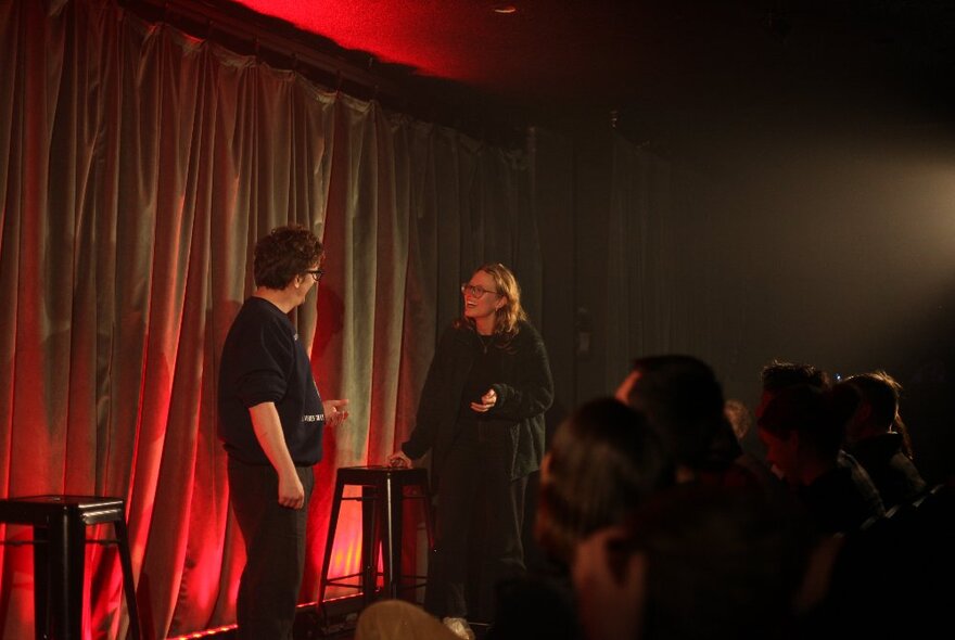 Two performers on a stage in front of a curtain, improvising a scene in front of an audience.