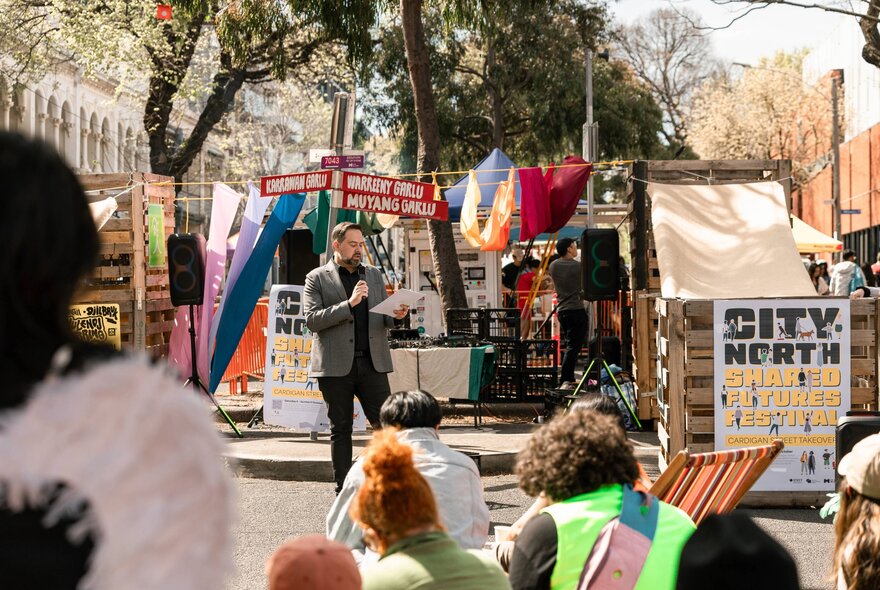 A person speaking into a microphone at an outdoor event, people looking on.