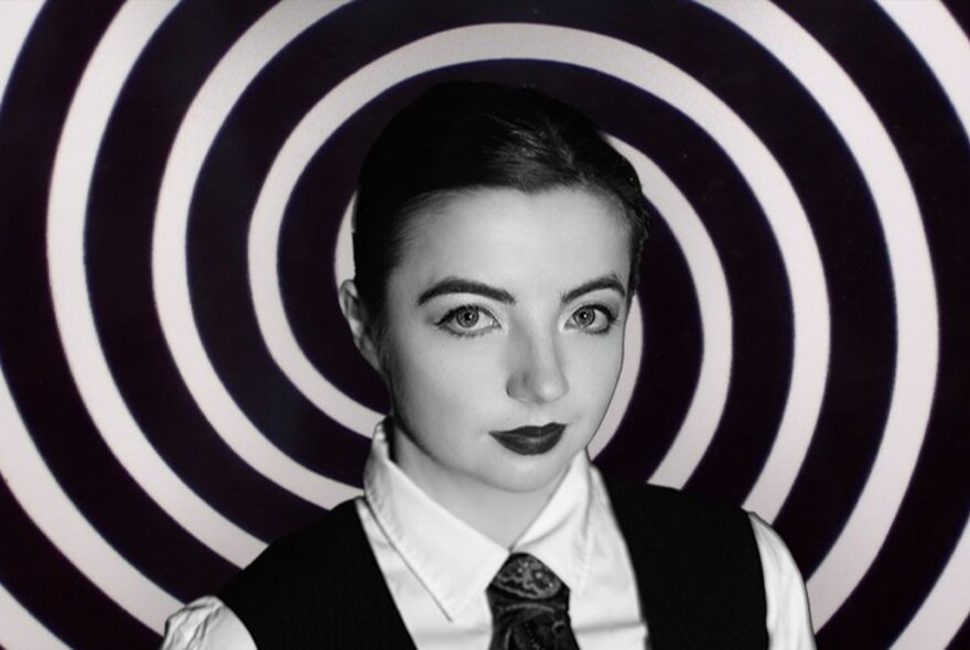 Woman wearing a tie and waistcoat with heavy makeup, black and white circles behind her.