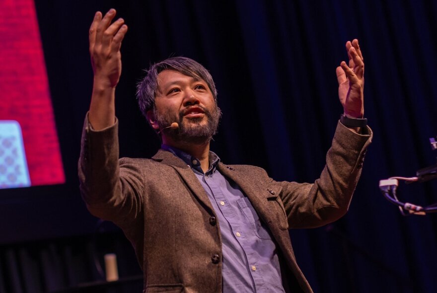 A man on stage with his arms raised, wearing a brown blazer and purple shirt.
