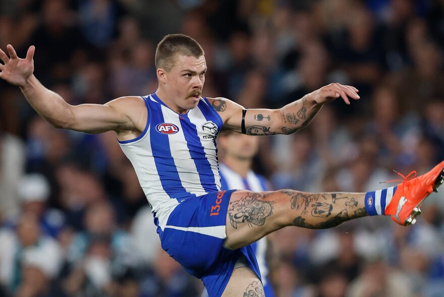 North Melbourne AFL football player kicking a ball during a game, his legs and arms tattooed.