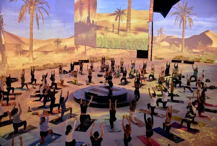 Wellness class seated exercising in a circle around a central dais with a backdrop of palm trees and dunes.