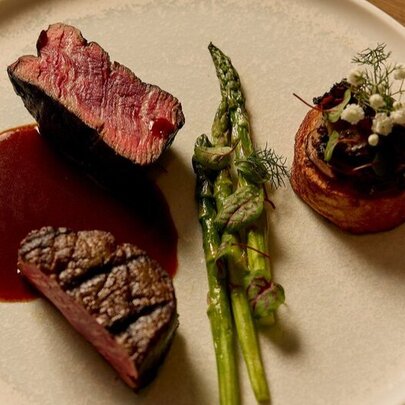 Looking down onto a white dinner plate with a slice of medium rare beef fillet resting on a sauce, with green asparagus spears.