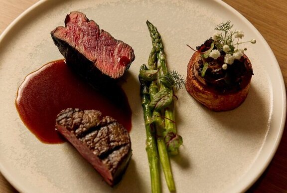 Looking down onto a white dinner plate with a slice of medium rare beef fillet resting on a sauce, with green asparagus spears.
